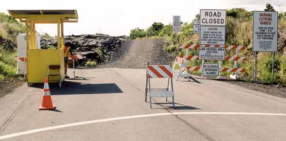 Booth and portable toilet at end of pavement, with one-lane gravel road continuing past sawhorses; multiple signs, including 'Road Closed', 'Lava viewing area | Open from | 12:00pm | Entry prohibited after 8:45pm | Gate will be locked after 10:00pm', 'Caution | Secure windows and | doors of your vehicle | during your visit here | Do not leave possessions unattended', and other signs shown earlier, with additional signs in background